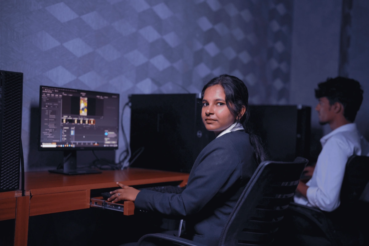 A woman in a suit sits at a computer desk, looking at the camera. Another person works at a computer in the background. The room is dimly lit.