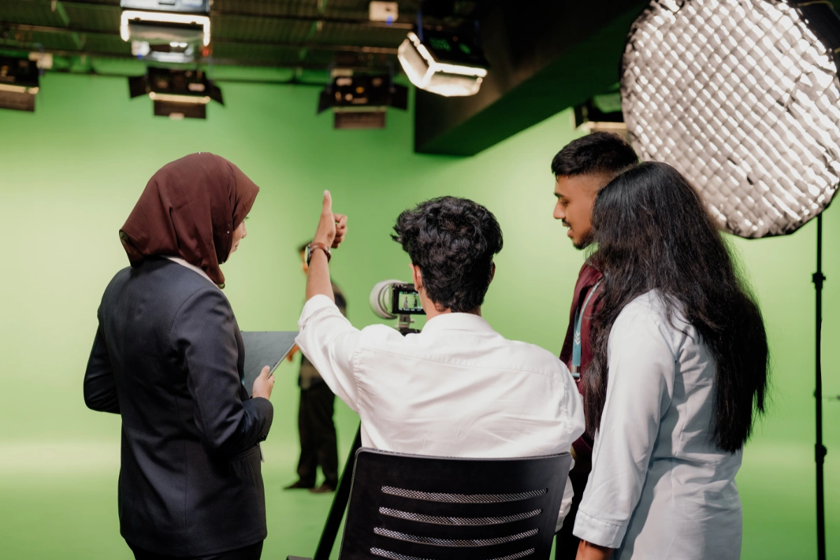 A diverse group of four people collaborate in a studio with a green screen backdrop. They are engaged and focused, surrounded by lights and filming equipment.