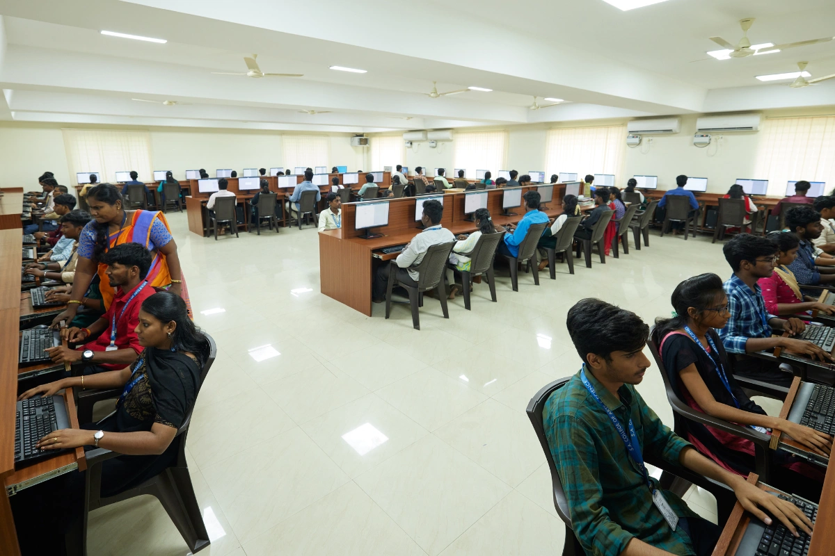 A spacious computer lab with rows of students working intently at desktop computers. The room is bright, organized, and focused on learning.