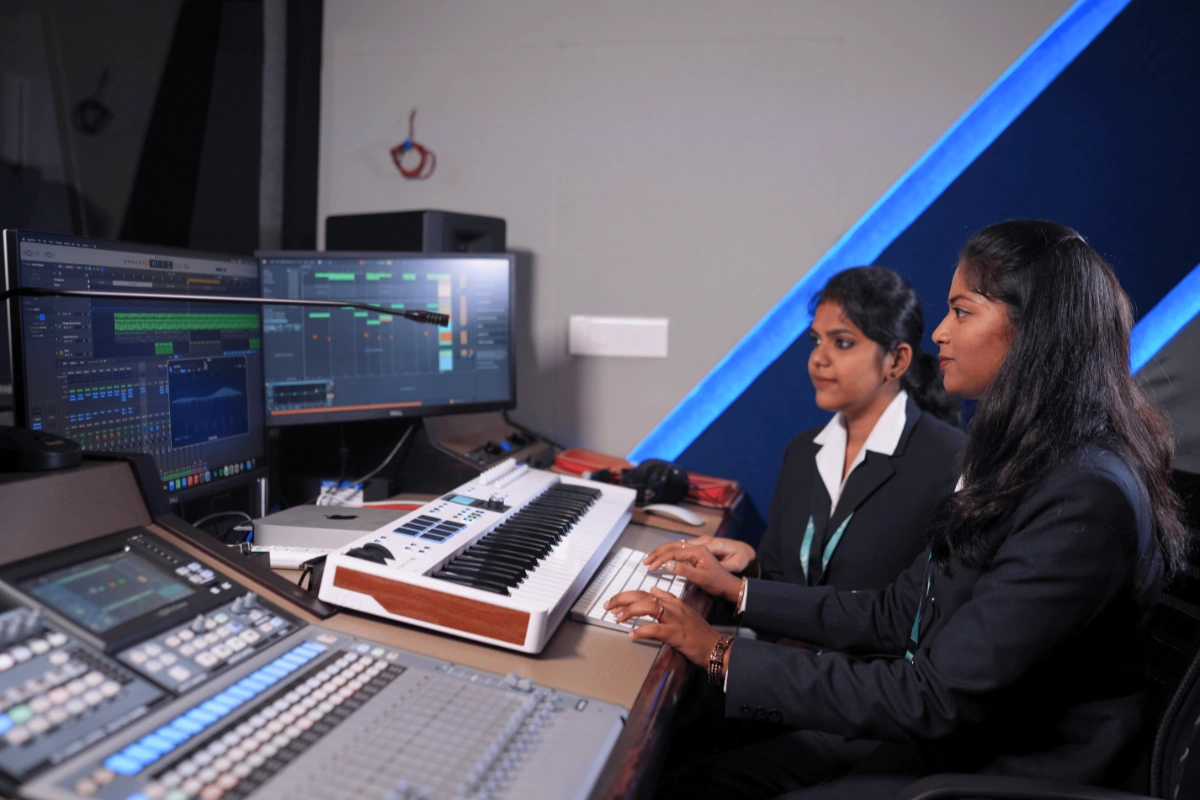Two women in a recording studio work at a computer with dual screens displaying audio editing software, alongside a keyboard and mixer.