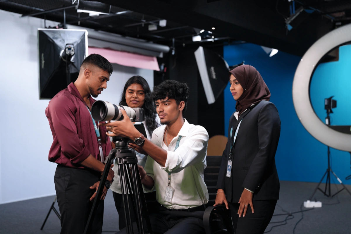 Four people in a studio setting work with camera equipment. One adjusts the camera on a tripod, while others watch attentively. The scene is professional and focused.